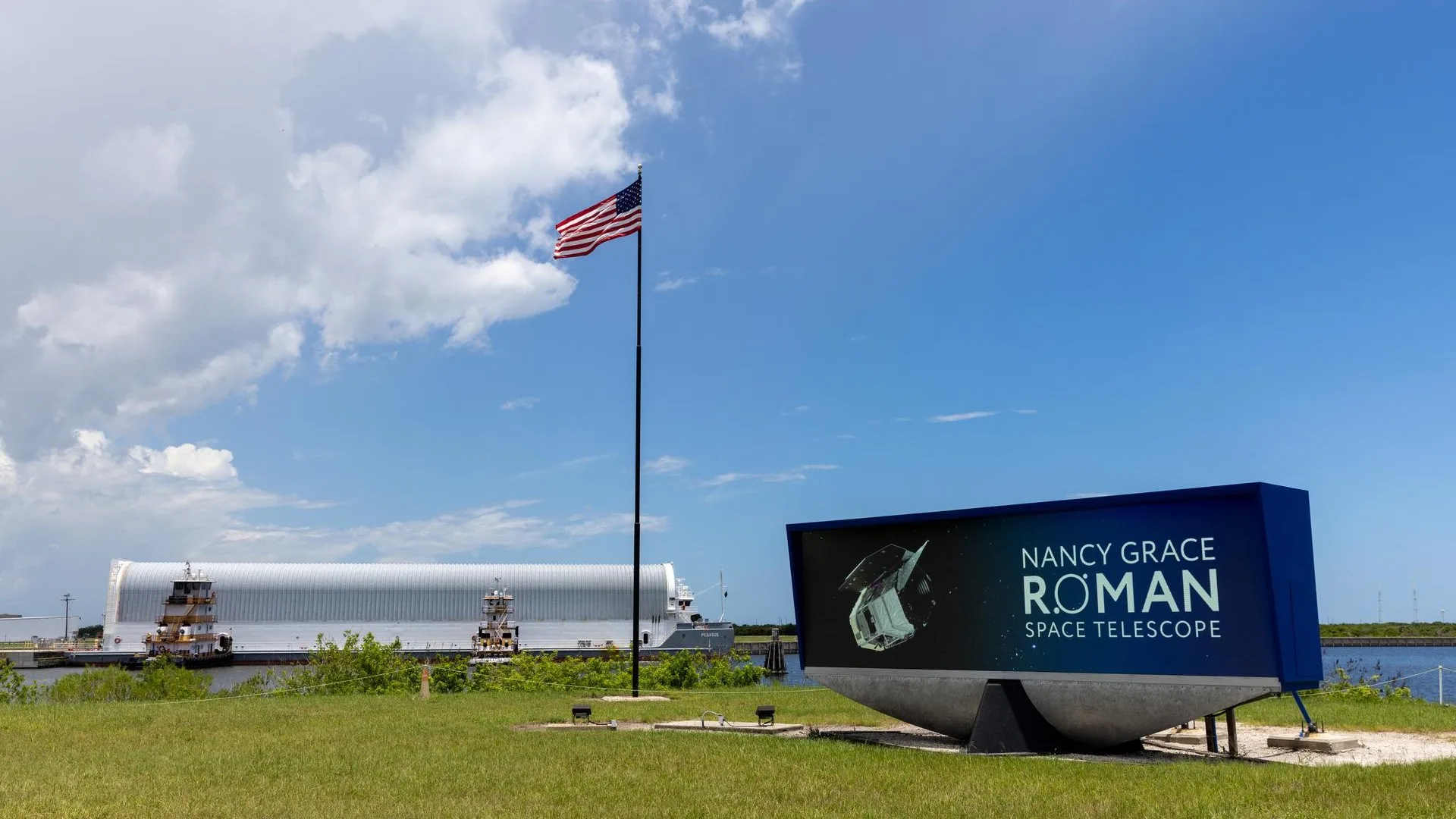 Roman Space Telescope Arrives in Florida for Major Falcon Heavy Launch Roman Space Telescope 2026 — a long barge in the background arrives at a small offloading dock. In the foreground, and A