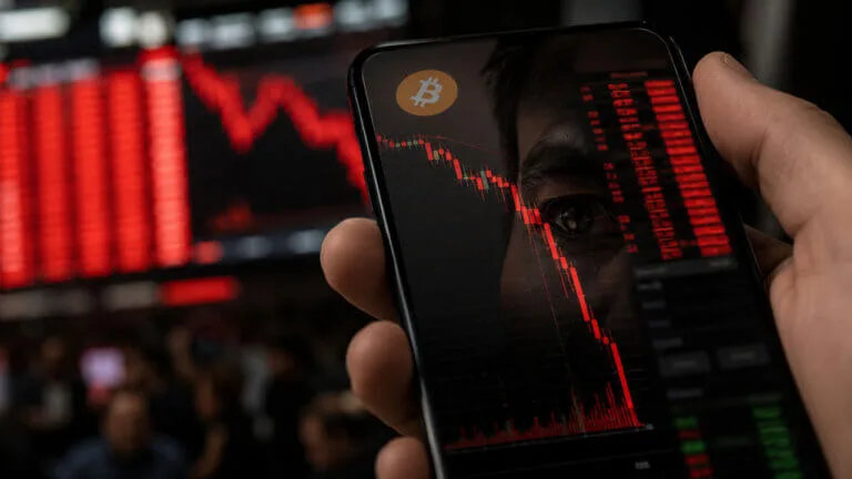 Bitcoin Price at $60K: Cheap on Chain, But Buyers Still Hesitate Close-up of a smartphone displaying a falling Bitcoin price chart, overlaid on a red market backdrop with a Bitcoin symb