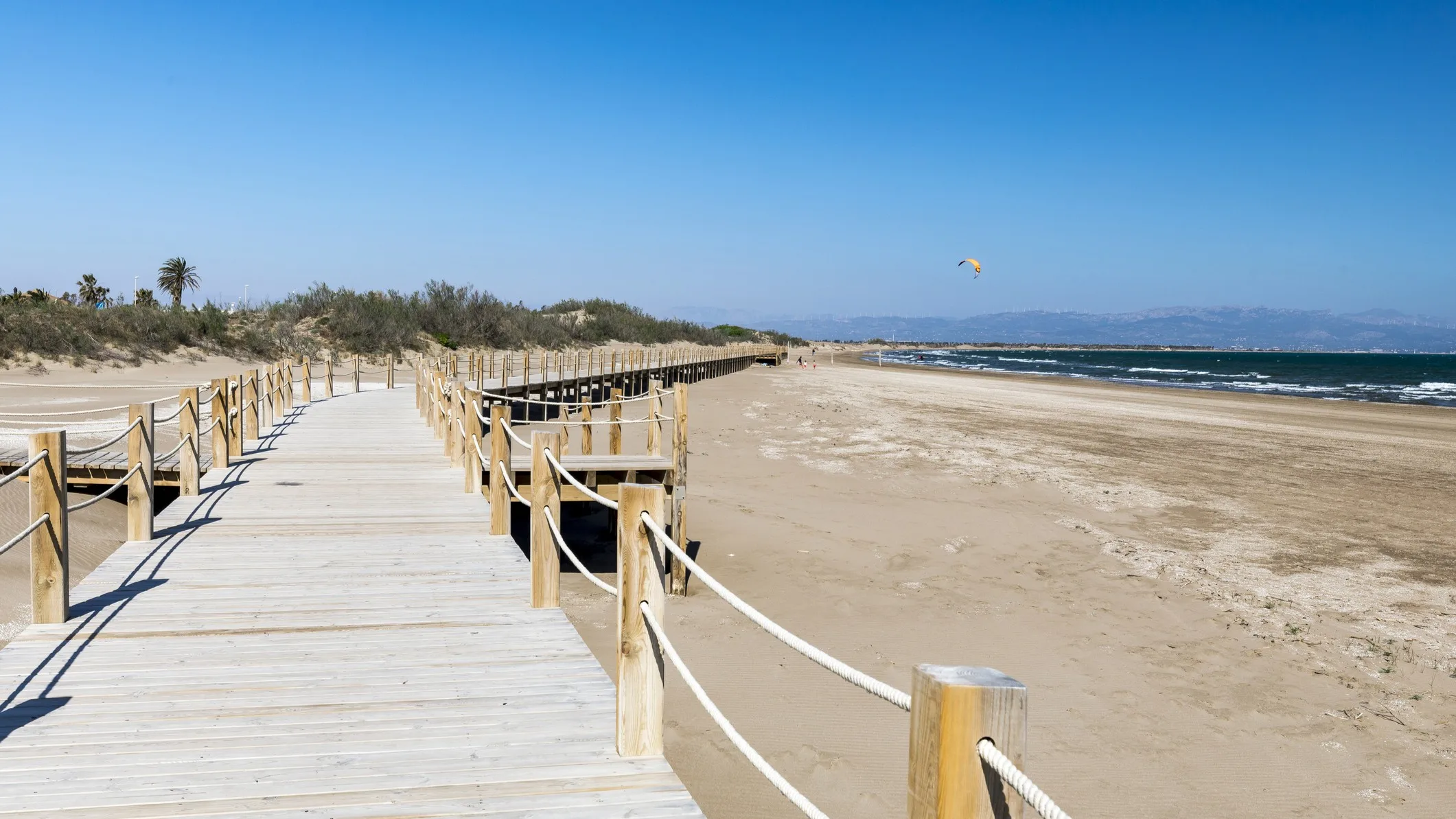 Best Spanish Beaches for the 2026 Total Solar Eclipse: 10 Top Picks sandy beach with a wooden boardwalk stretching into the distance. The sky and ocean are a deep blue.