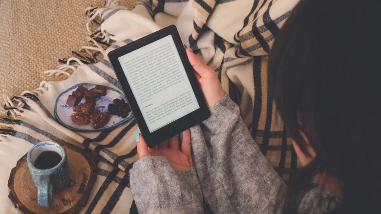 Free Kindle Books: The Best Sources You're Not Using Yet A woman sitting on a blanket reads from her Kindle, sampling a cup of coffee and a plate of treats.
