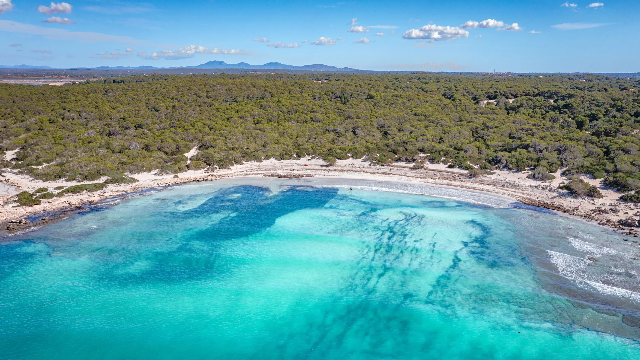 Best Spanish Beaches for the 2026 Total Solar Eclipse: 10 Top Picks aerial view of a white sand beach and azure blue waters and sky above. Behind the sanding beach is vast swathes of green