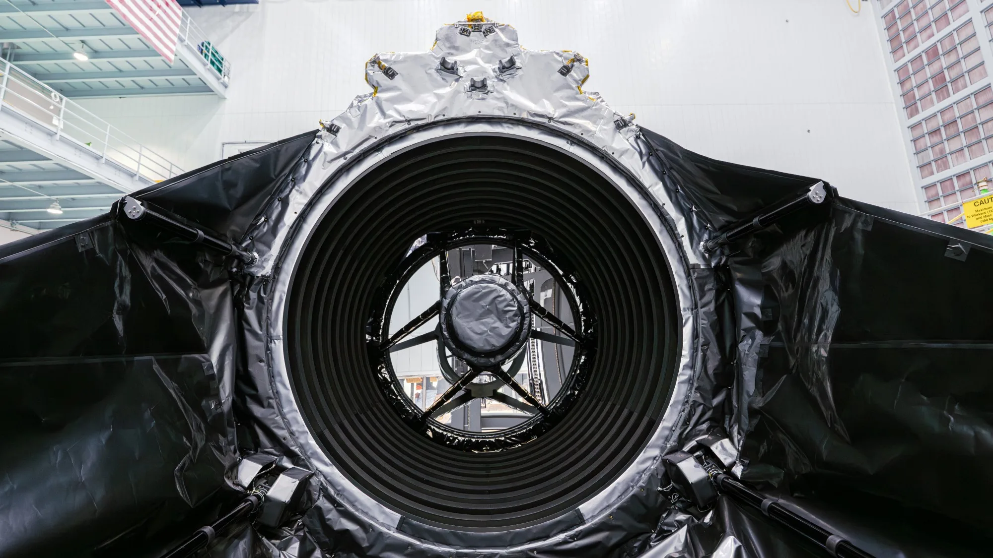 NASA Roman Space Telescope Passes Final Mirror Check Before Launch technicians in clean suits in a hangar next to a large rectangular spacecraft wrapped in metallic foil