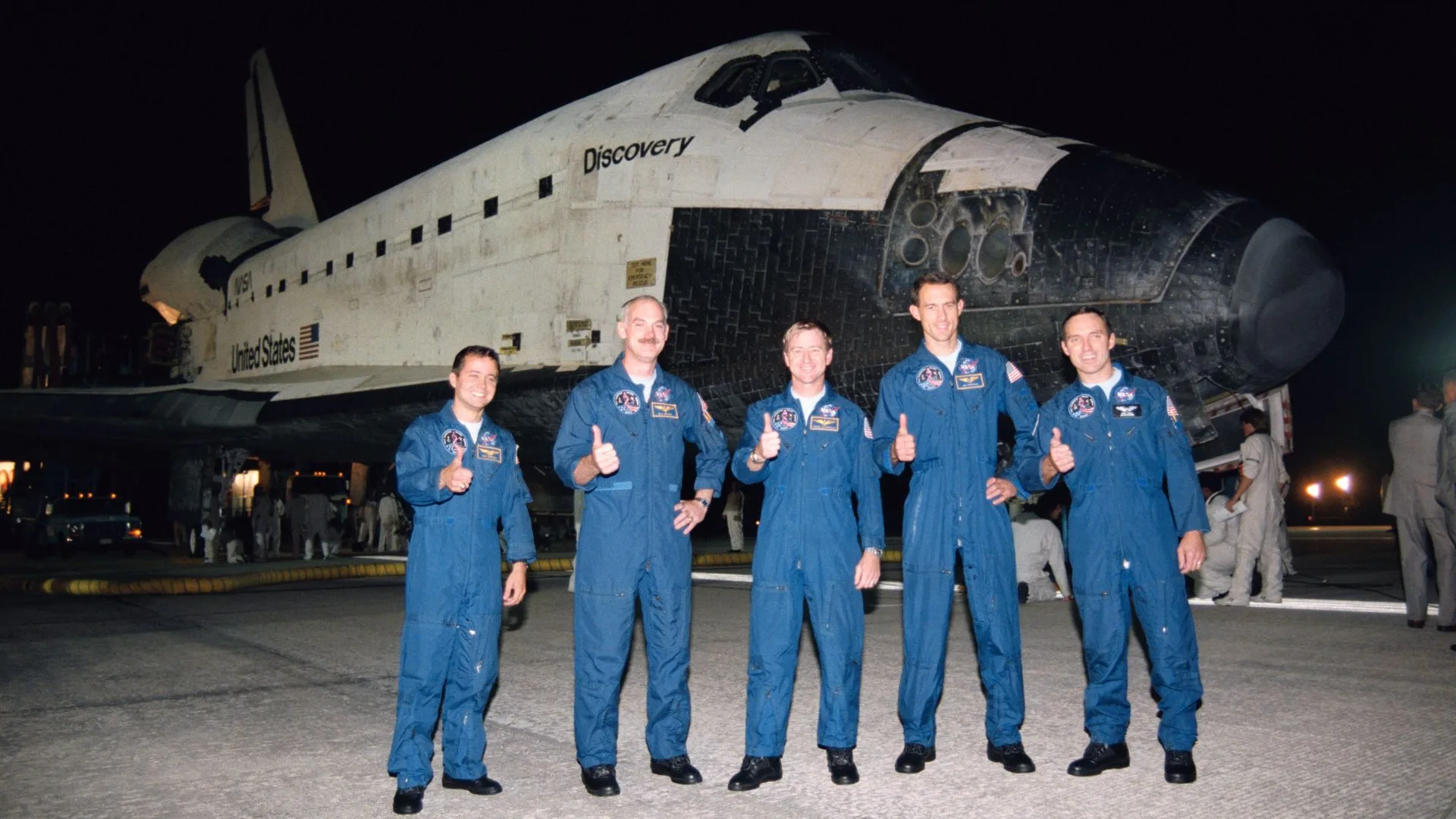 Moon Missions and Meteor Storms: The Critical Risk NASA Is Planning Fo A crew of five astronauts in blue flightsuits stand giving the thumbs up in front of a space shuttle at night