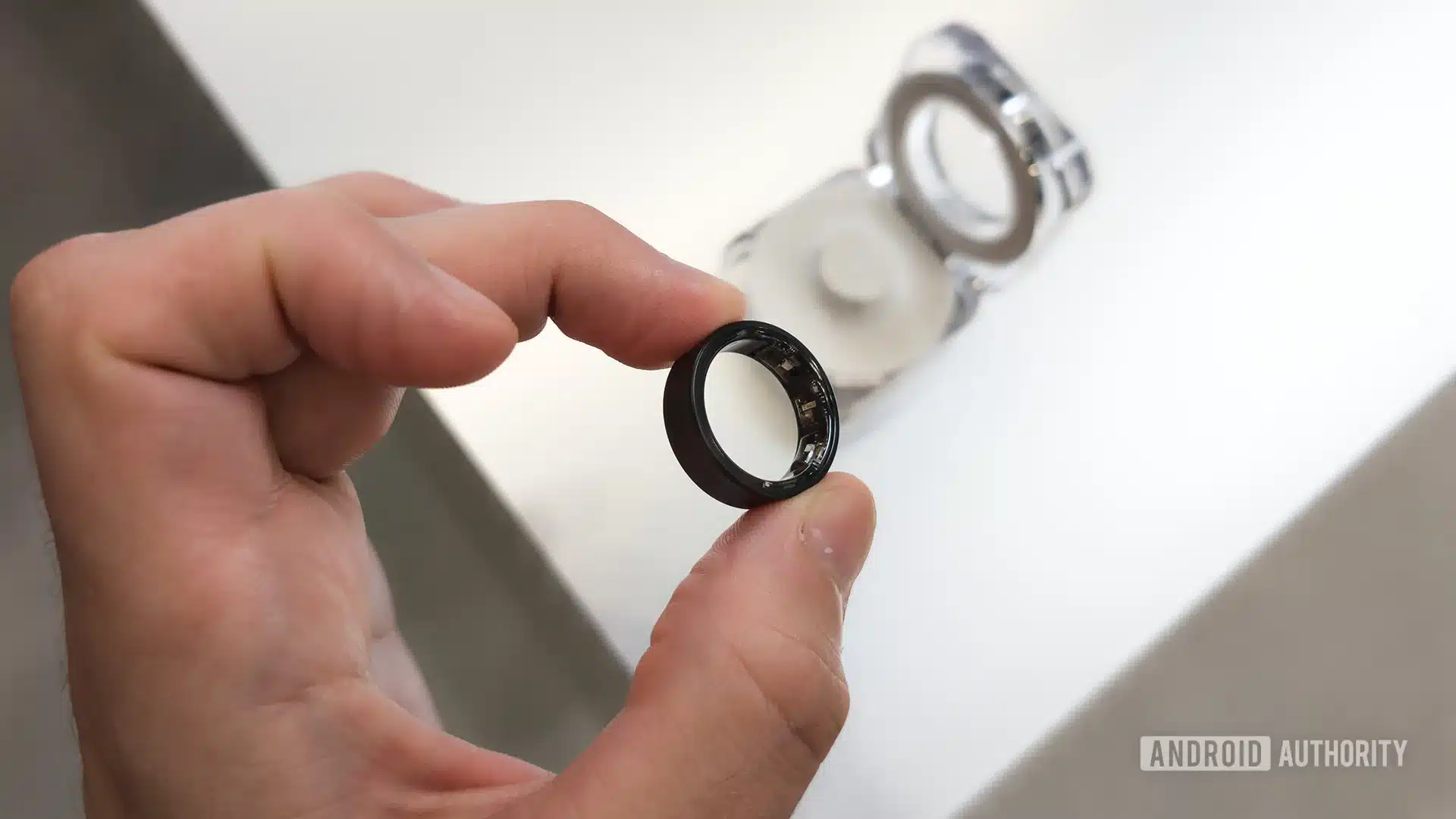 Galaxy Ring 2 Confirmed — and It May Support iPhone Galaxy Ring 2 — Samsung Galaxy Ring in Titanium Black Held Between Two Fingers with Case in Background