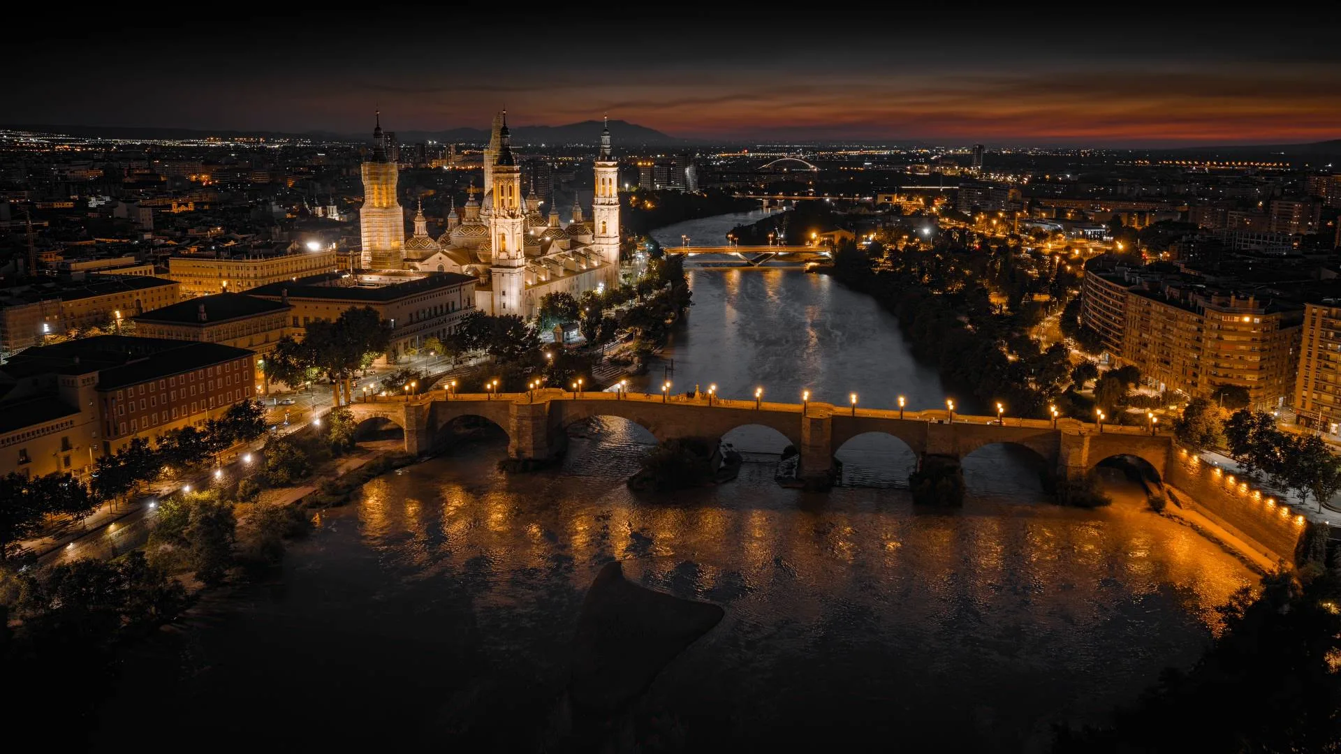 Spain Solar Eclipse 2026: Top 10 Cities for Best Views A cityscape at dusk featuring a brightly lit cathedral, stone bridge, and river reflecting golden lights, with buildings