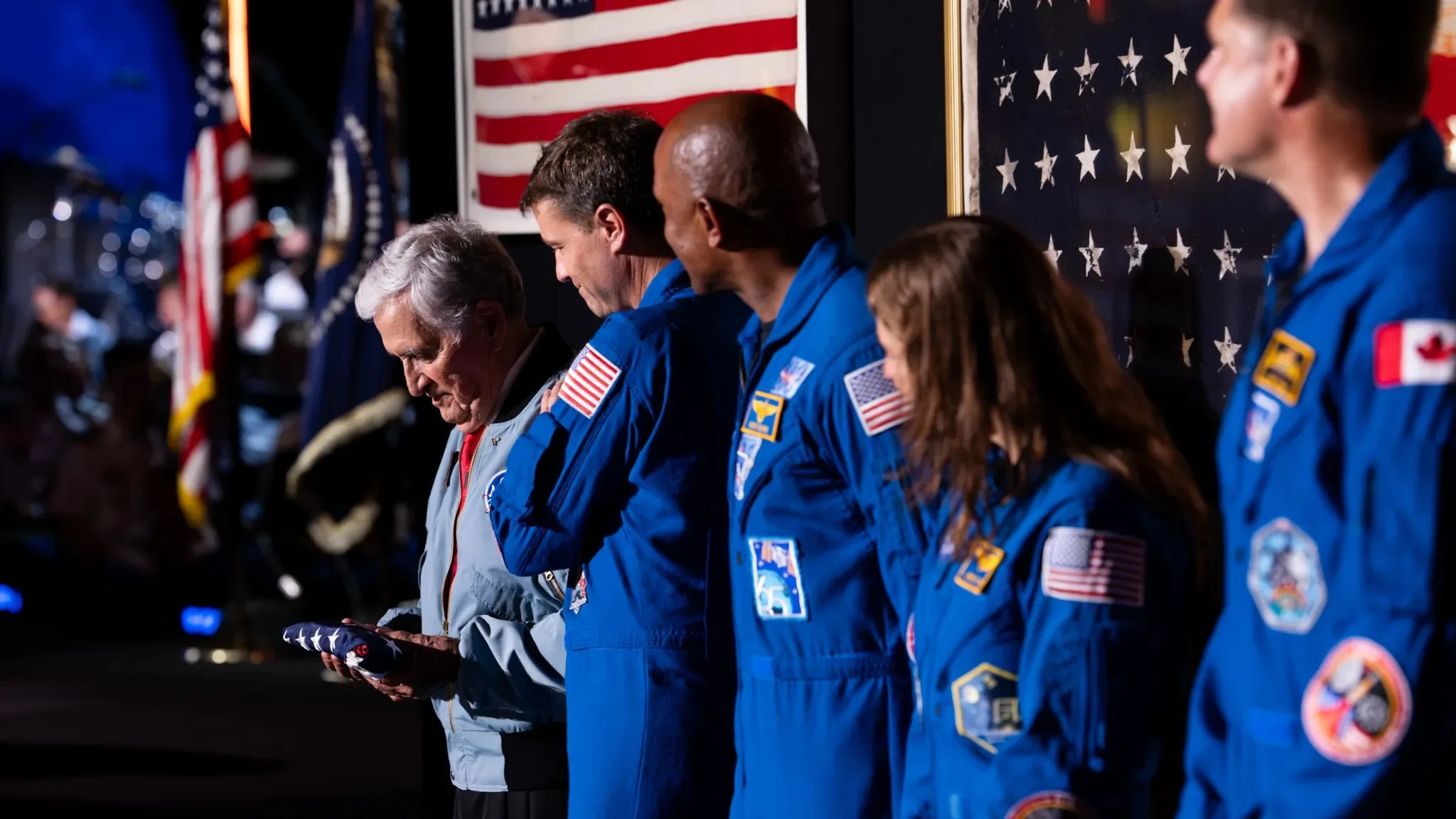 Artemis Moon Mission Gets an Official Flag — Handed Over by Apollo 17' Artemis moon mission — four people in blue jumpsuits from the right, look left and a grey-haired man in a light blue jac