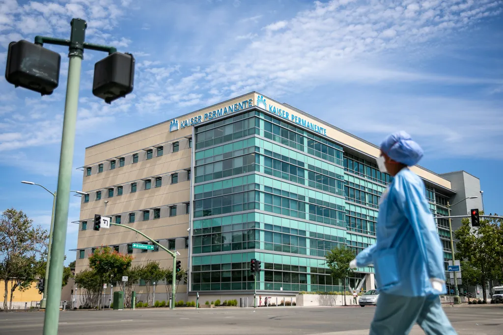 Kaiser Nurse Surveillance Is a Critical Test for Healthcare AI A person dressed head to toe in blue scrubs looks towards a Kaiser Permanente as they walk across an intersection.