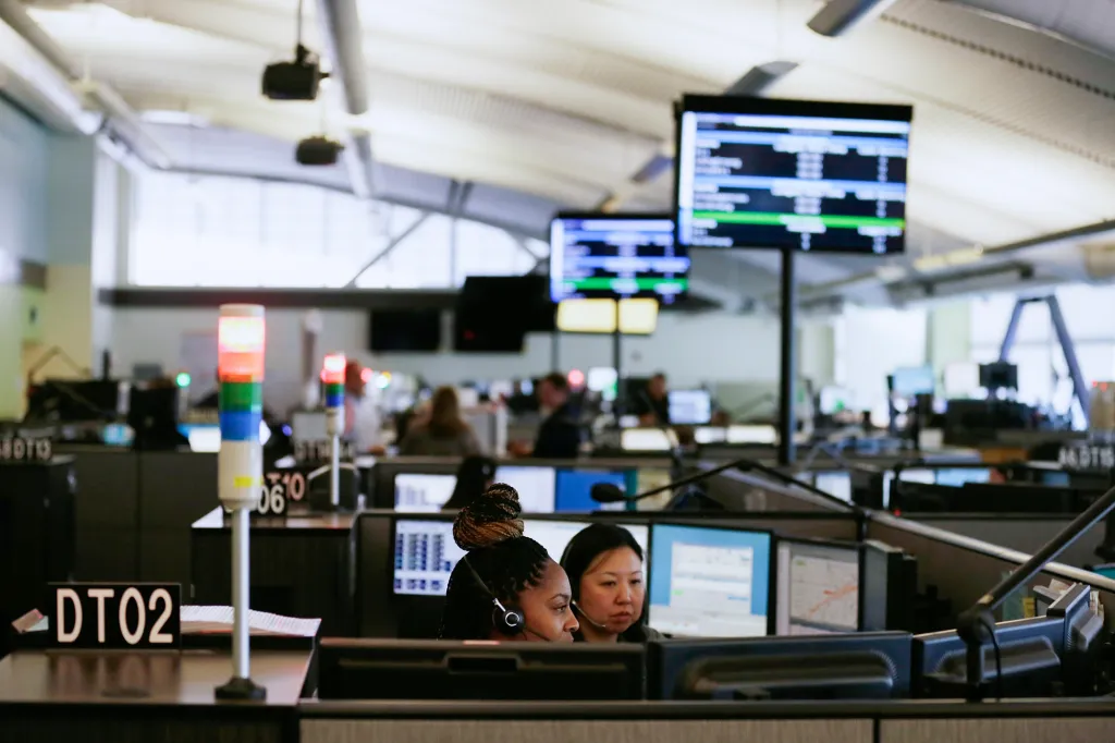 Kaiser Nurse Surveillance Is a Critical Test for Healthcare AI A wide view inside a 911 dispatch center with rows of computer stations and overhead screens displaying call and dispatc