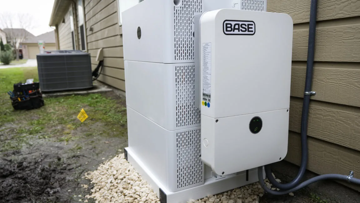 Home Battery Installations Hit Record 673 MW in Early 2026 A white-colored residential battery with the name "BASE" on the side sits on the side of a brown-colored house